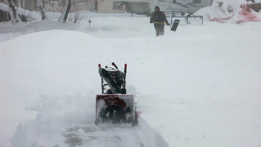Man walking through deep snow with a shovel towards a snow blower stuck in deep snow blizzard. Snow blowing in strong wind in sub freezing weather. Winter time hazard and climate nature.