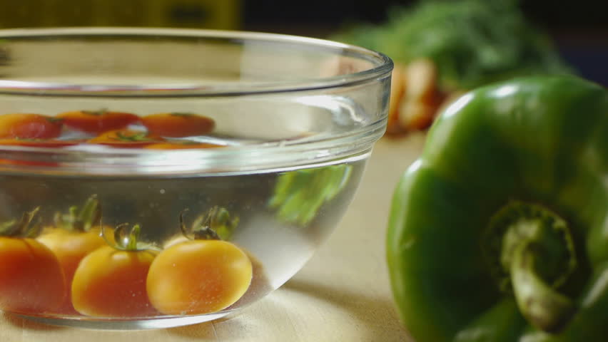 red tomatoes and broccoli being washed in bowl of water, with peppers and other vegetables on kitchen table. Dolly shot