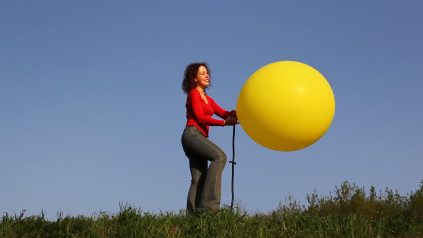 young woman stands in meadow with green grass against blue sky and blows up yellow balloon with foot pump  