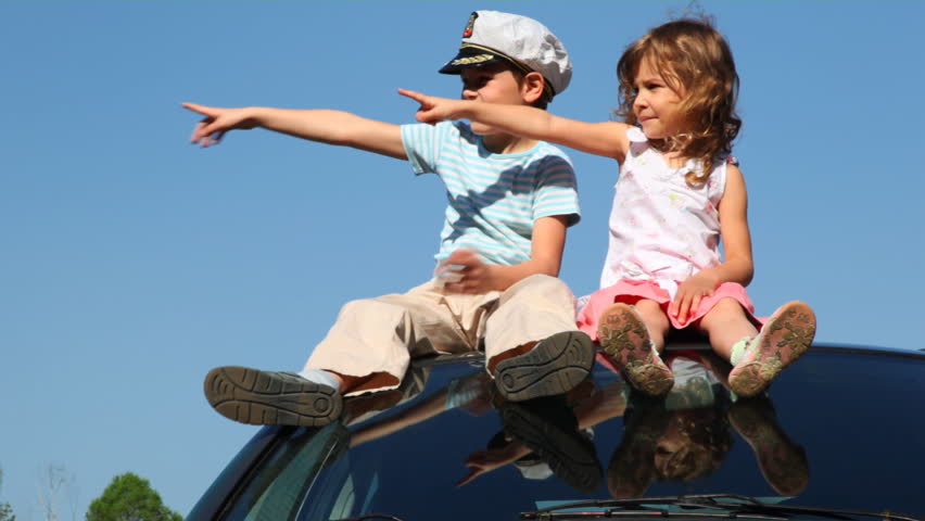 boy in captain's cap and his little sister sit on roof of car against blue sky pointing somewhere with their hands, then twist imaginary driving wheel