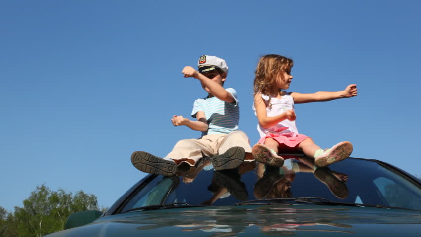 boy in captain's cap and his little sister sit on roof of car against blue sky spinning imaginary driving wheel