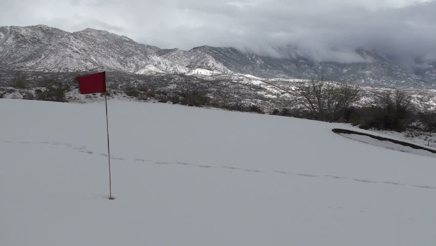 Red flag flutters in snowstorm that blankets golf course white, against backdrop of beautiful, snow-covered mountains. 1920x1080