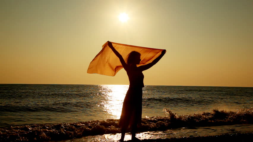 pretty girl stands on beach and holds over her head scarf fluttering in breeze, silhouette at sunset evening