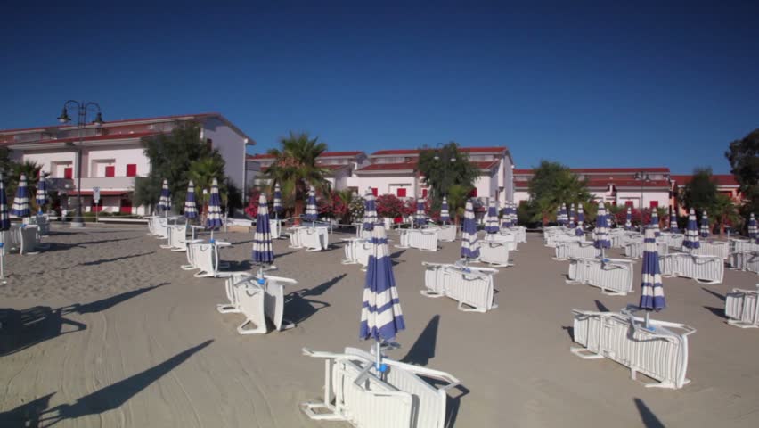 Many parasols and deckchairs standing on beach at sunny day, some house is at background, camera moving
