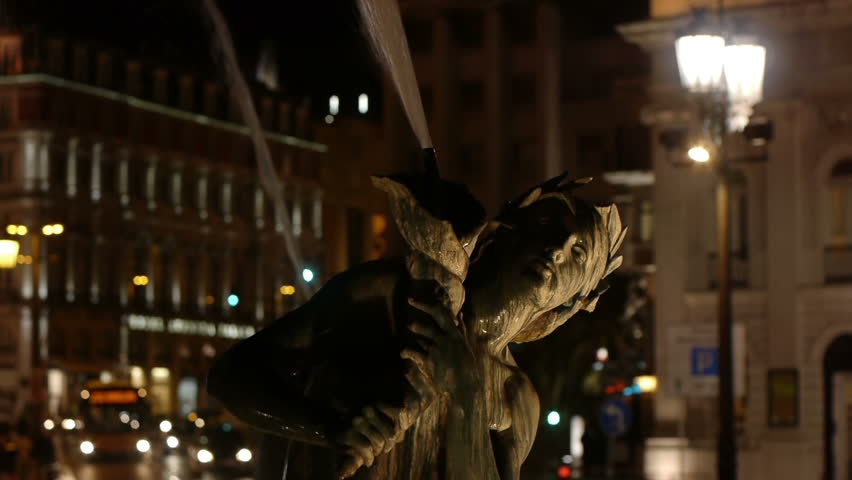 Night time video of a fountain statue with cars and people passing in the background. Version with lamp on the upper right visible.