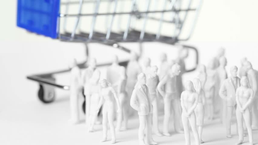 Group of little colorless toy men and women stand in front of big shopping trolley, all composition rotates
