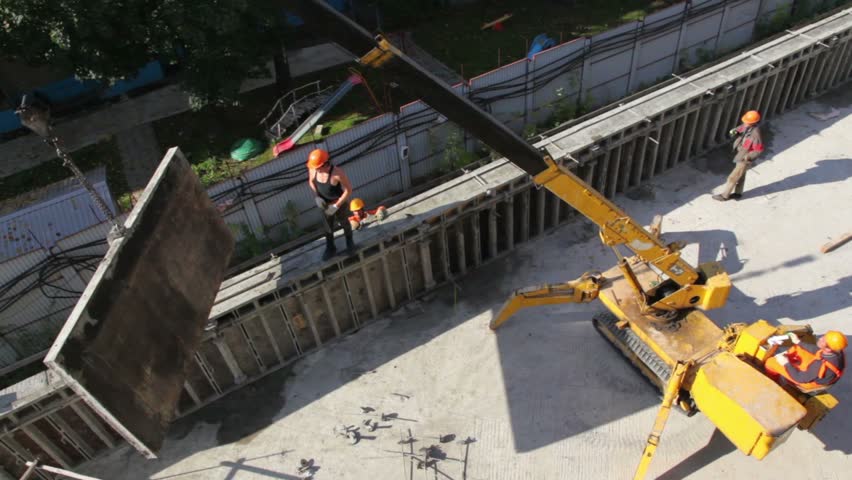 Small crane lifts part of wall and carries it, several workers are around