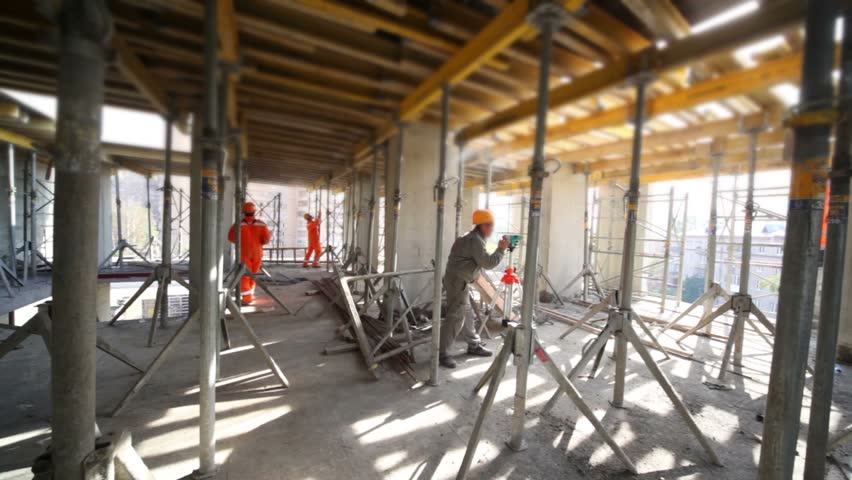 Several builder work in unfinished premises, one uses leveling instrument, camera moves and shows room around