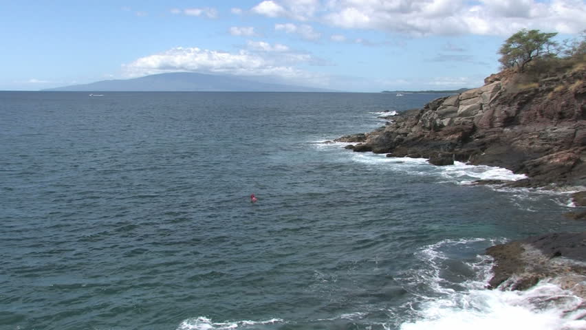 rocky shoreline on the island of Maui in Hawaii. A man is swimming and spear fishing in the rough water. Beautiful water with cliff and trees overlooking water. Island of Lanai on horizon.