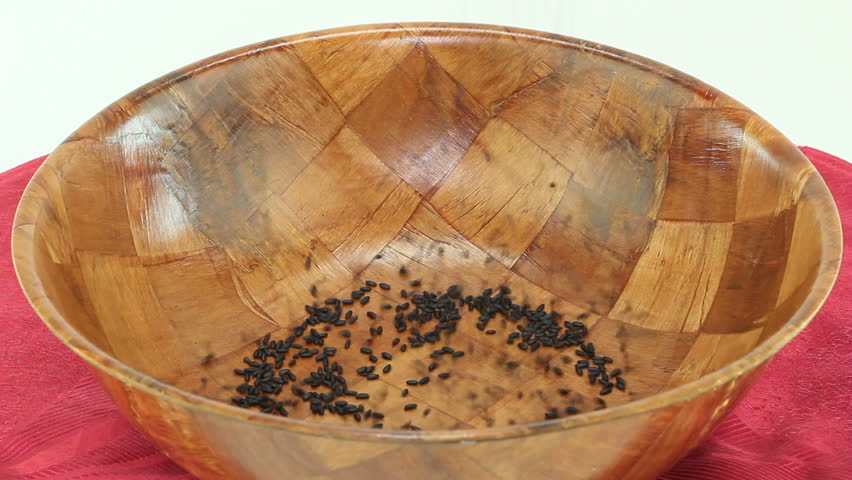 Dry purple rice that looks black falls into a round wood bowl on a red placemat in front of a white background.