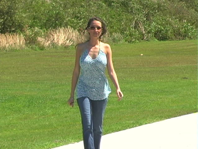 A beautiful brunette walks toward the camera down a wide sidewalk on a breezy summer day.