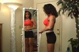 A pretty brunette with remarkable muscle definition, dressed in fitness wear, measures her waist in front of a full-length mirror.