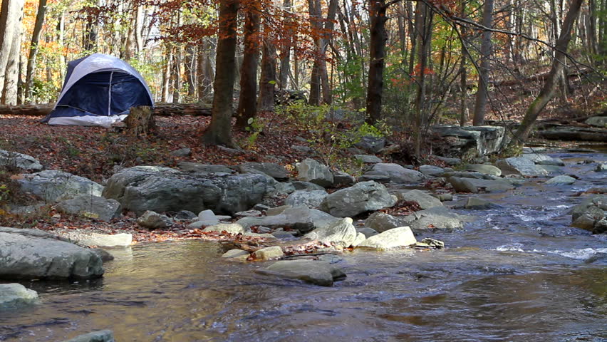 Small camping tent is pitched by a mountain stream in the woods in autumn with fall foliage.