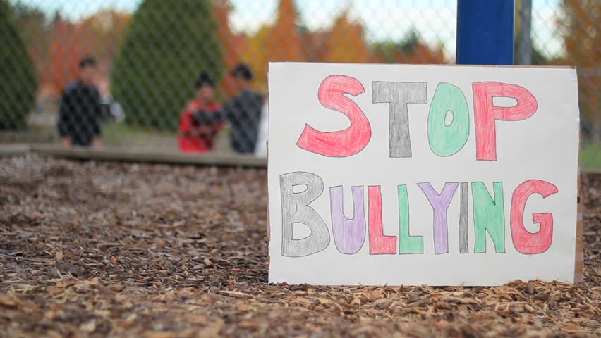 'Stop Bullying' sign. In the background, a group of teenagers push, shove and bully a minority student on the basketball court at school.