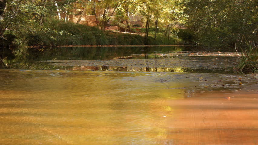 The waters of Oak Creek in Sedona, Arizona ripple slowly over a concrete area as the fall leaves are reflected in the early morning and sunlight runs across the surface.