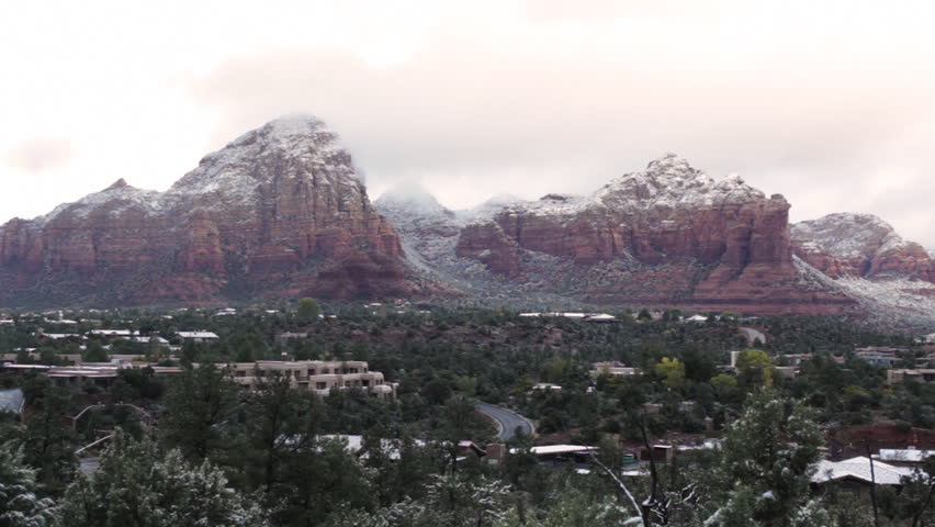 Sunlight shines down through heavy, early-morning clouds covering the mountains of Sedona, Arizona - Coffee Pot Rock - with a light dusting of snow. Wide shot. Juniper trees in foreground.