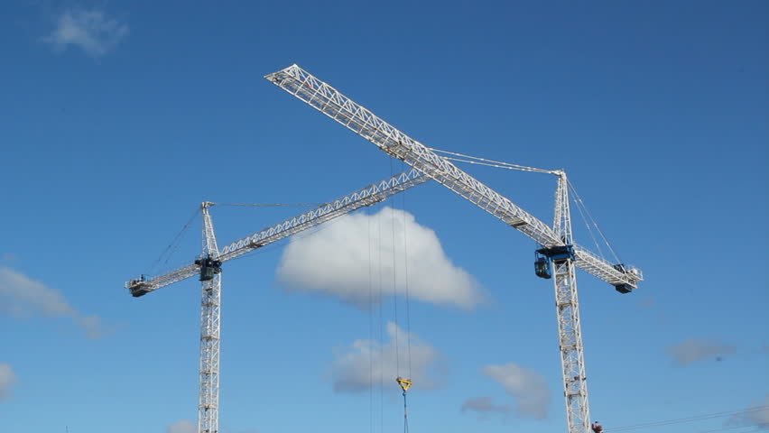 Two cranes.  White tower cranes against blue sky. Trolley moves along jib of foreground crane. Realtime shot. Construction in Toronto. 