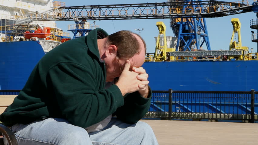 Depressed unemployed man sits on a bench at the shipyard with his head in his hands.
