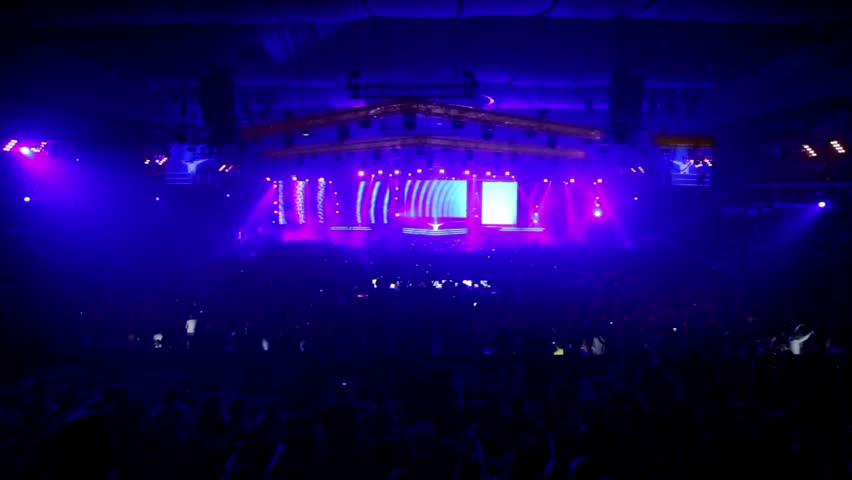Lot of fans in large stadium at rave party, dj  rises hands on stage, bright light flashes