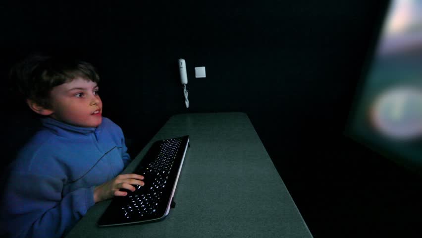 Young boy chews gum and plays video game with keyboard on big screen in dark room