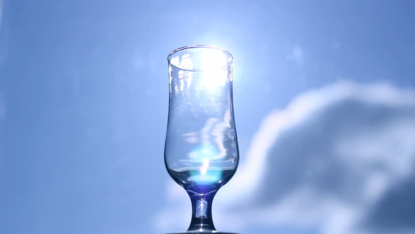 Pouring water from a bottle into a glass, with a sun and blue sky in background. HD1080i. 