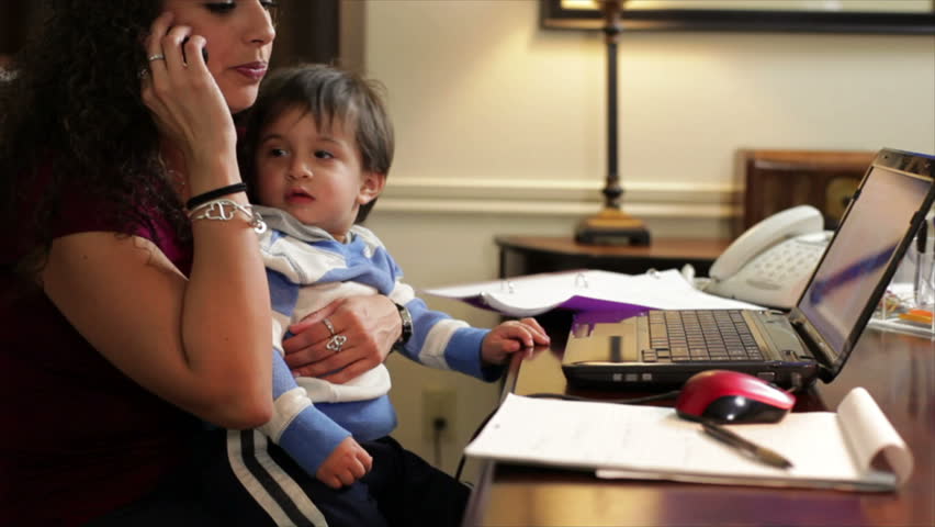 A pretty Hispanic work-at-home mom holds her son while taking a phone call and making notes on a writing table.