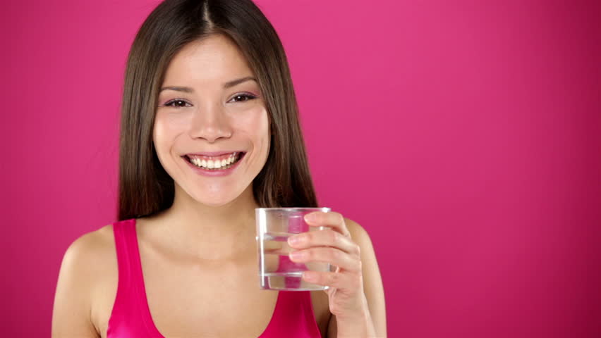 Woman drinking water. Happy smiling girl drinking glass of water smiling looking at camera and side on pink background. Beautiful young mixed race Asian / Caucasian female model.