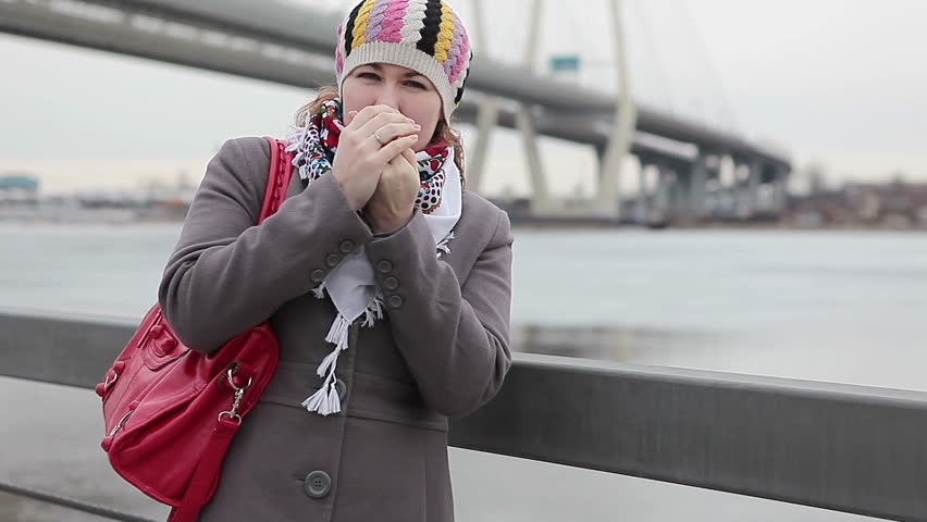 Frozen young Caucasian female warming hands and standing on embankment. Bridge on background 