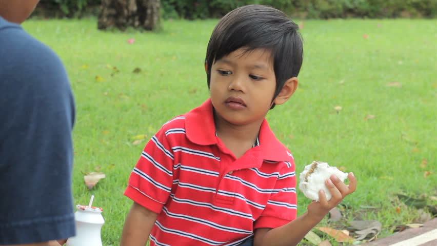A loving father gives his cute Asian boy a drink of milk while having a picnic lunch at the park.