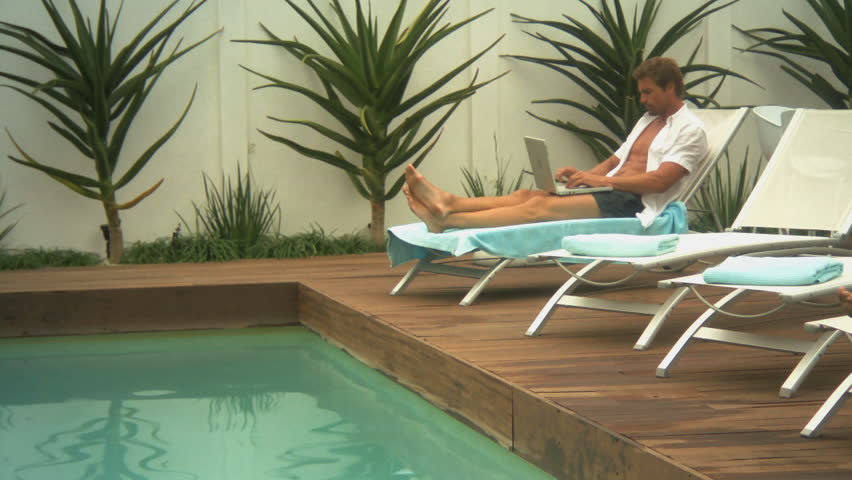 LONG SHOT PAN OF A MAN WORKING ON A LAPTOP AND THEN DIPPING HIS FEET IN A SWIMMING POOL