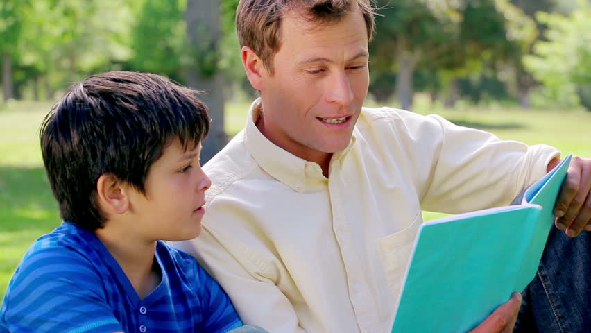 Smiling man reading a book with his son in a parkland