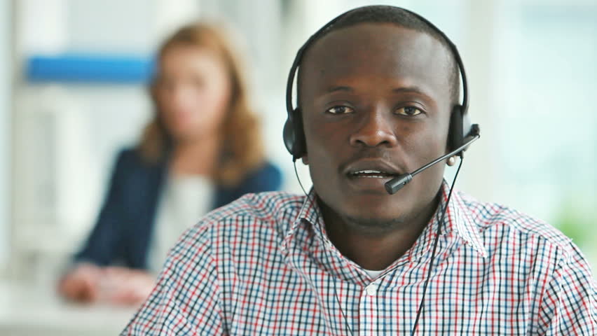 African-American man wearing headset answering calls of clients, his colleague can be vaguely seen in the background