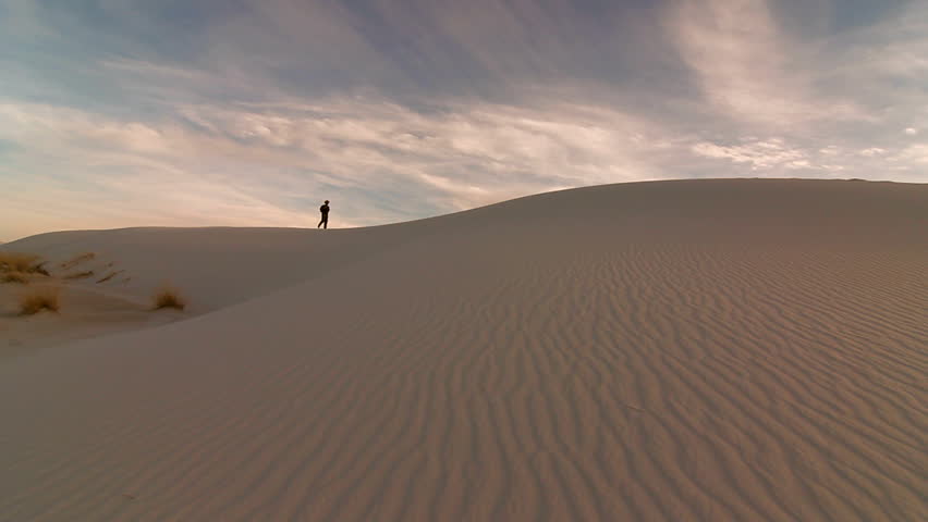 Lone silhouetted hiker walks across sand dunes beneath dramatic cloud-streaked sky. 1080p