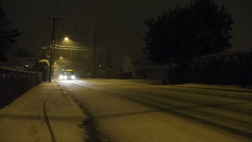Fresh snow falls at night in Portland, Oregon as traffic including city busses and small car drive cautiously on roads.