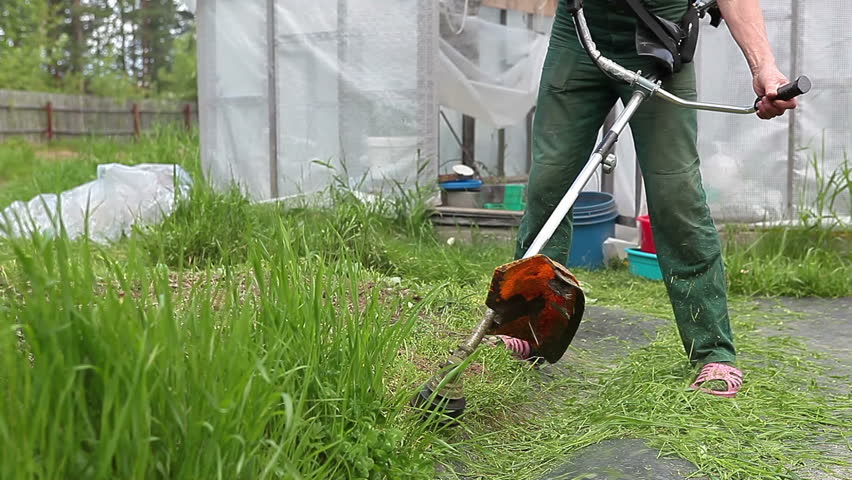 Unrecognizable male mows overgrown green grass by petrol trimmer 