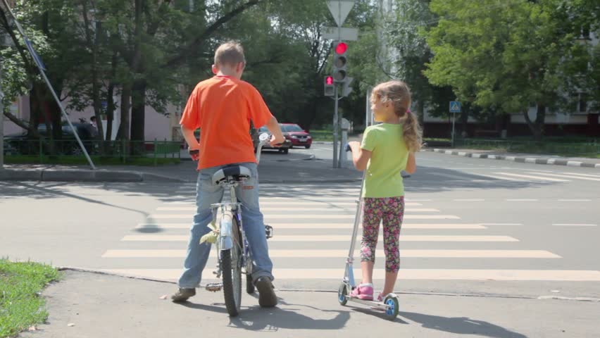 Boy sits on bicycle and his sister stands with scooter at pedestrian crossing