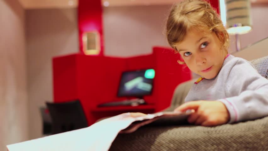 Little girl sits in chair and reads aloud newspaper at room with computer on desk