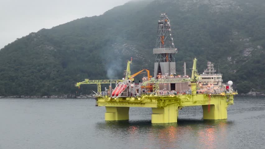 Oil rig located in sea near coastline with forest on mountain under cloudy sky