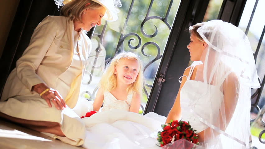 Cute little blonde girl dressed flower girl laughing with grandmother and older sister in wedding dress