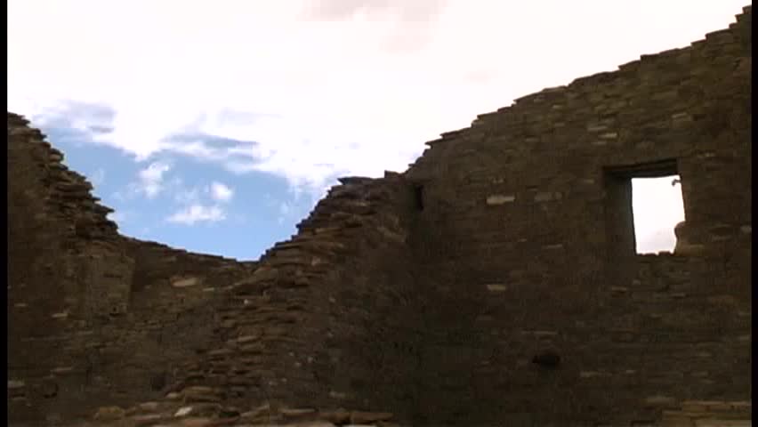 A black raven glides over the fallen walls of Pueblo Bonito in Chaco Canyon on a windy day, with blue, partly cloudy sky in the background