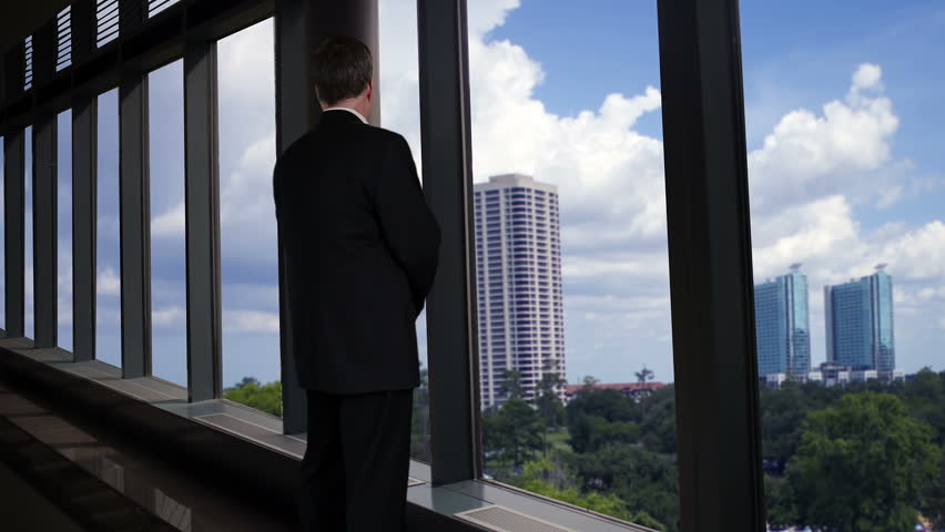 A businessman looking out the large plate glass windows of a long skywalk turns and smiles with satisfaction.