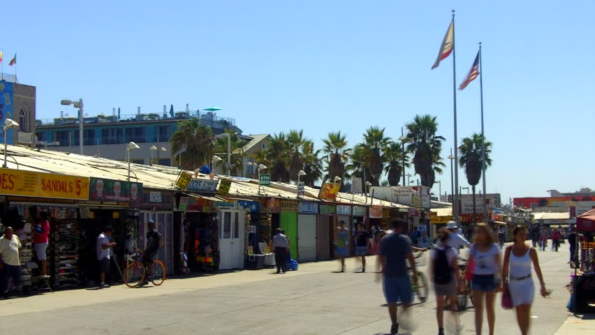 A time lapse shot of people walking, riding bikes, roller skating, riding skateboards at the boardwalk in Venice Beach, CA with souvenir stands in the background. 
