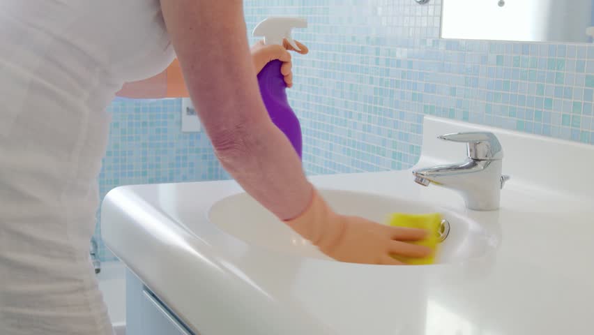 Senior woman doing chores in bathroom at home, cleaning sink and faucet with spray detergent. Sequence