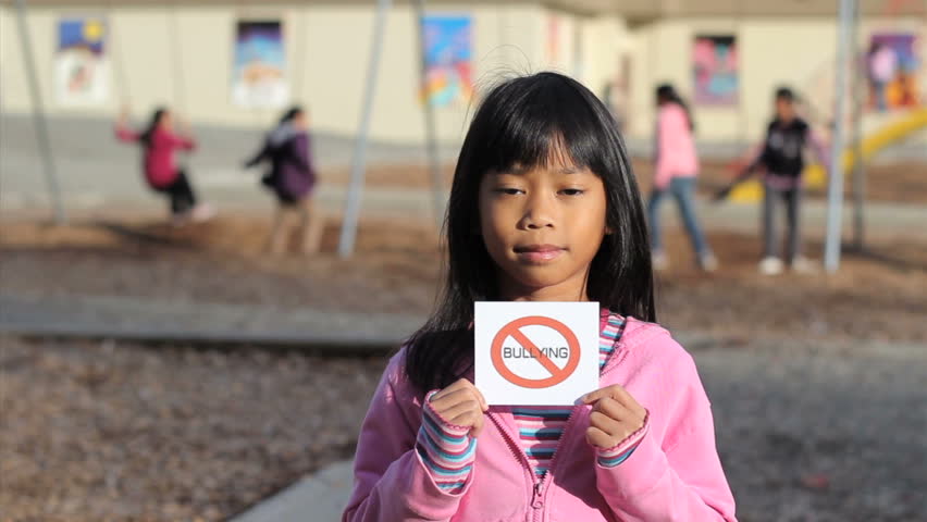 A cute Asian girl holds up a small NO BULLYING sign on the school playground.