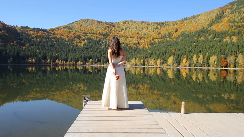 Lonely bride in wedding dress holding a small flower bouquet in hands, admiring the amazing nature scenery of the autumn colorful trees mirrored in the mountain lake