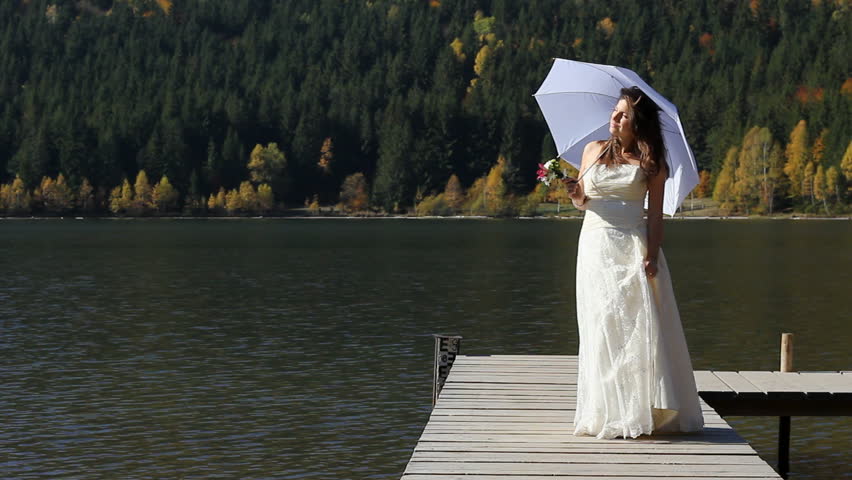 Single beautiful bride wearing white wedding dress and umbrella walking on a wooden pontoon and admiring the amazing scenery of the autumn forest mirrored in the mountain lake during a sunny day