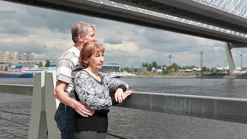 Loving mature couple under city bridge on river embankment looking around. Summer day