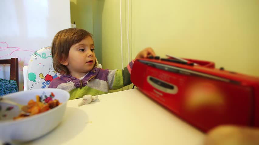 Girl toddler sits at table and plays with a red radio set. Early morning breakfast.
