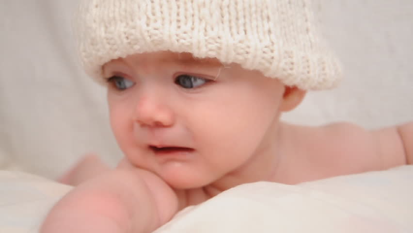A little baby is crying and sad wearing a white hat on a bed with a blanket.
