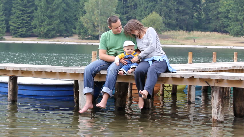 Young family, barefoot father, mother and baby sitting together and relaxing on a wooden pontoon at the shore of a mountain lake